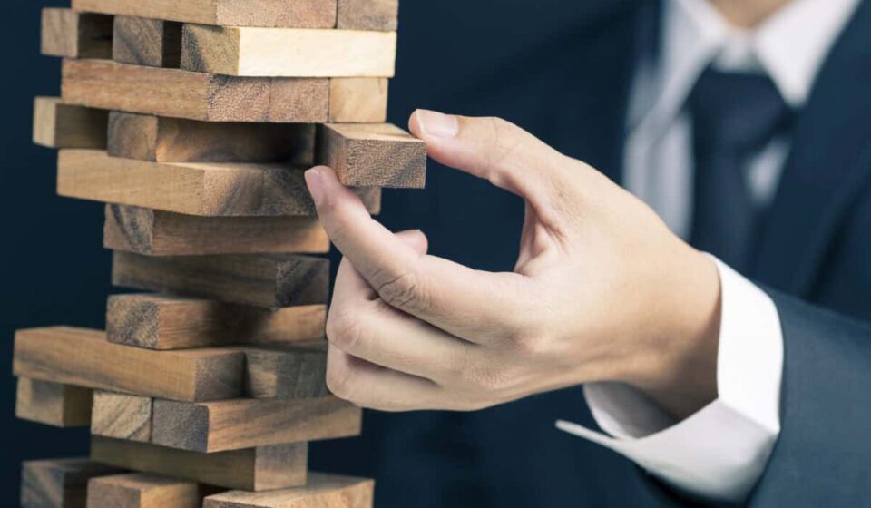 man in business suit pulls a piece out of wobbly wooden tower